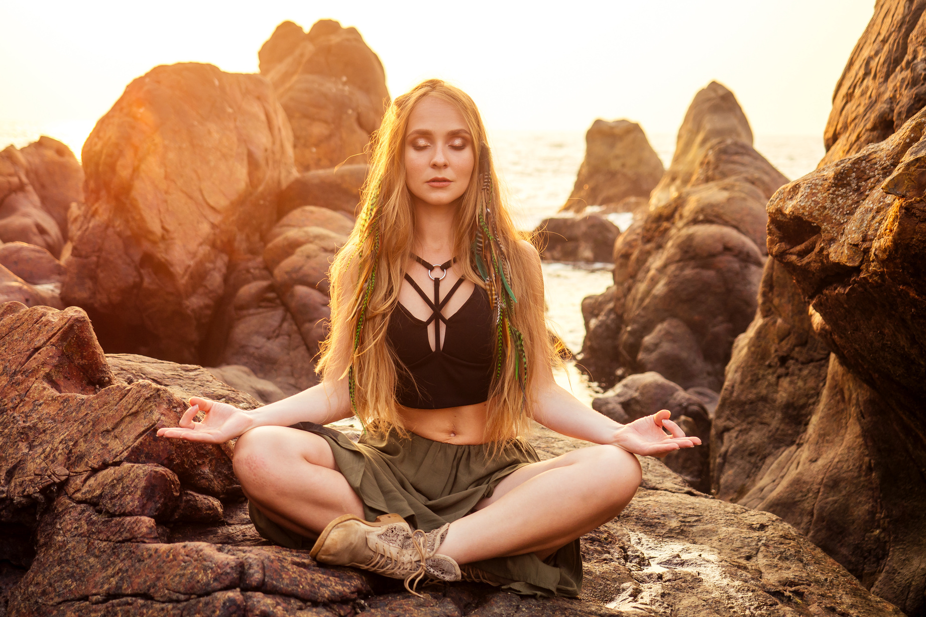 stylish redheaded chestnut blonde young girl in indie  bo-ho style clothes black stylish top. woman long hair,makeup and green feathers in head practicing yoga asanas on the rocks by sea